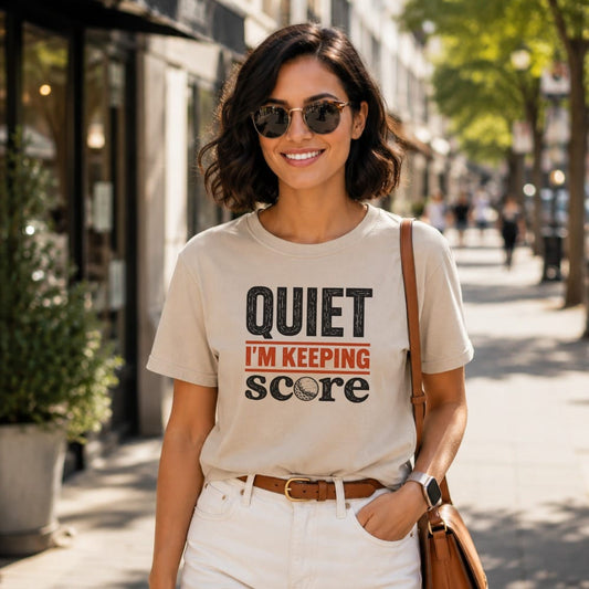 Woman wearing a tan t-shirt with "QUIET I'M KEEPING SCORE" text and golf ball graphic.