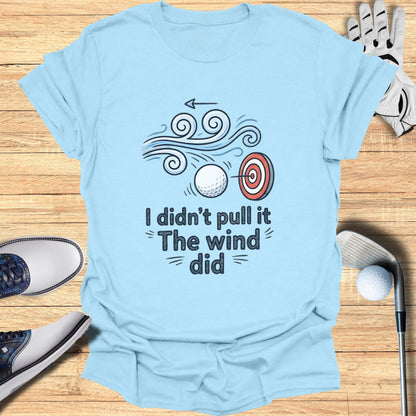 Light blue t-shirt with a graphic of a golf ball being blown by wind towards a target.