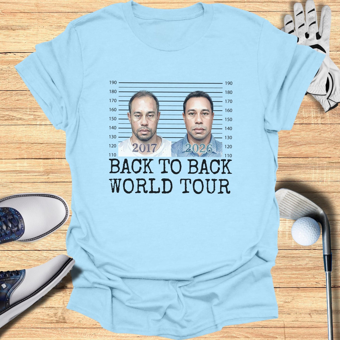 Light blue t-shirt with mugshot-style images of Tiger Woods from 2017 and 2026, text reads "BACK TO BACK WORLD TOUR".