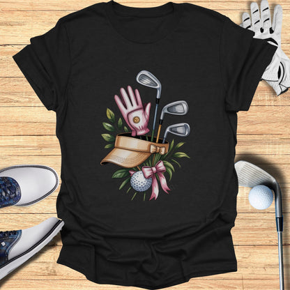 Golf clubs, visor, glove, ball, and pink ribbon on black t-shirt.