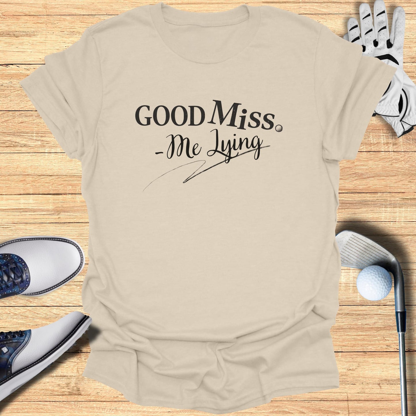 Light tan t-shirt with "Good Miss -Me Lying" text, golf accessories.