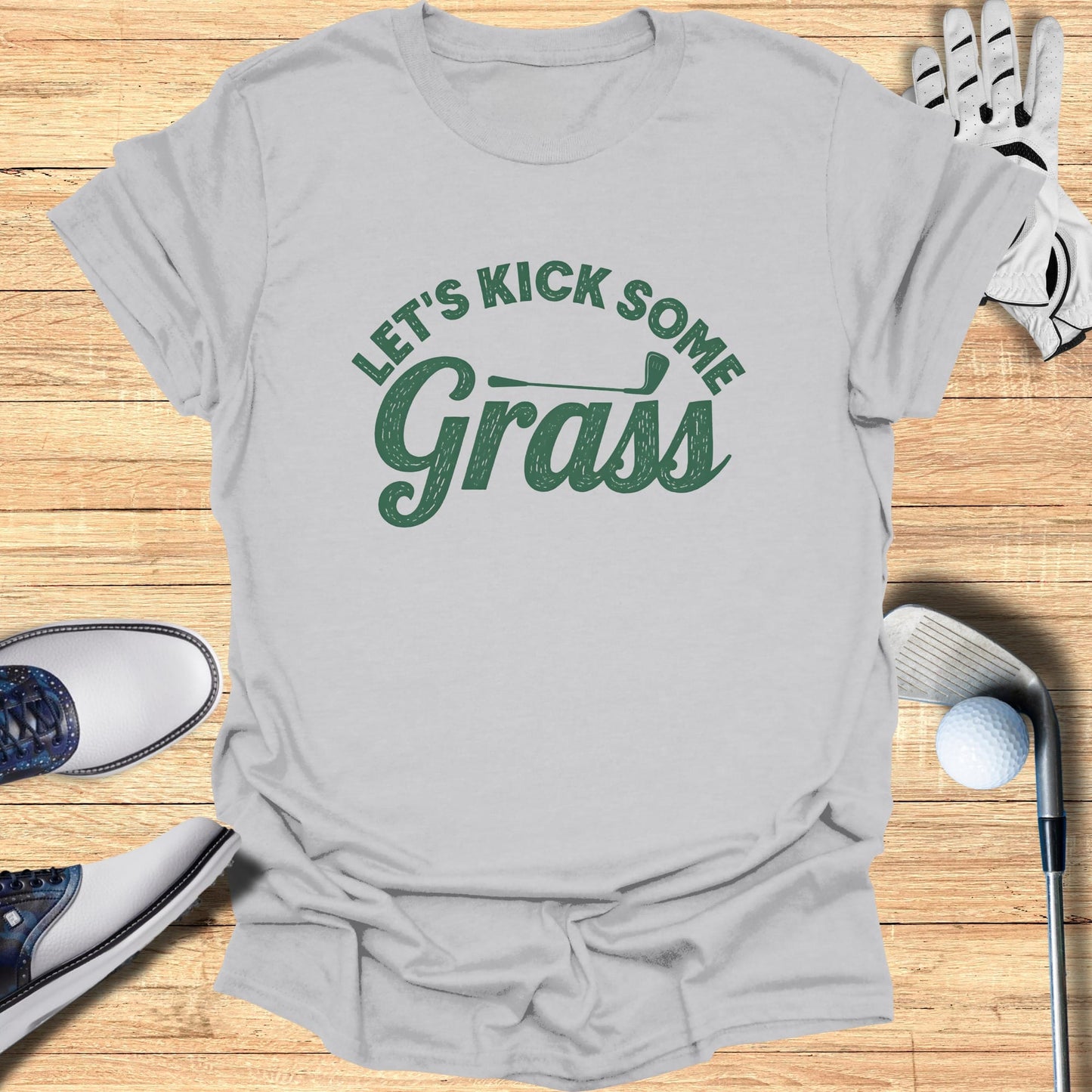 Gray t-shirt with green text "Let's Kick Some Grass" above golf accessories.