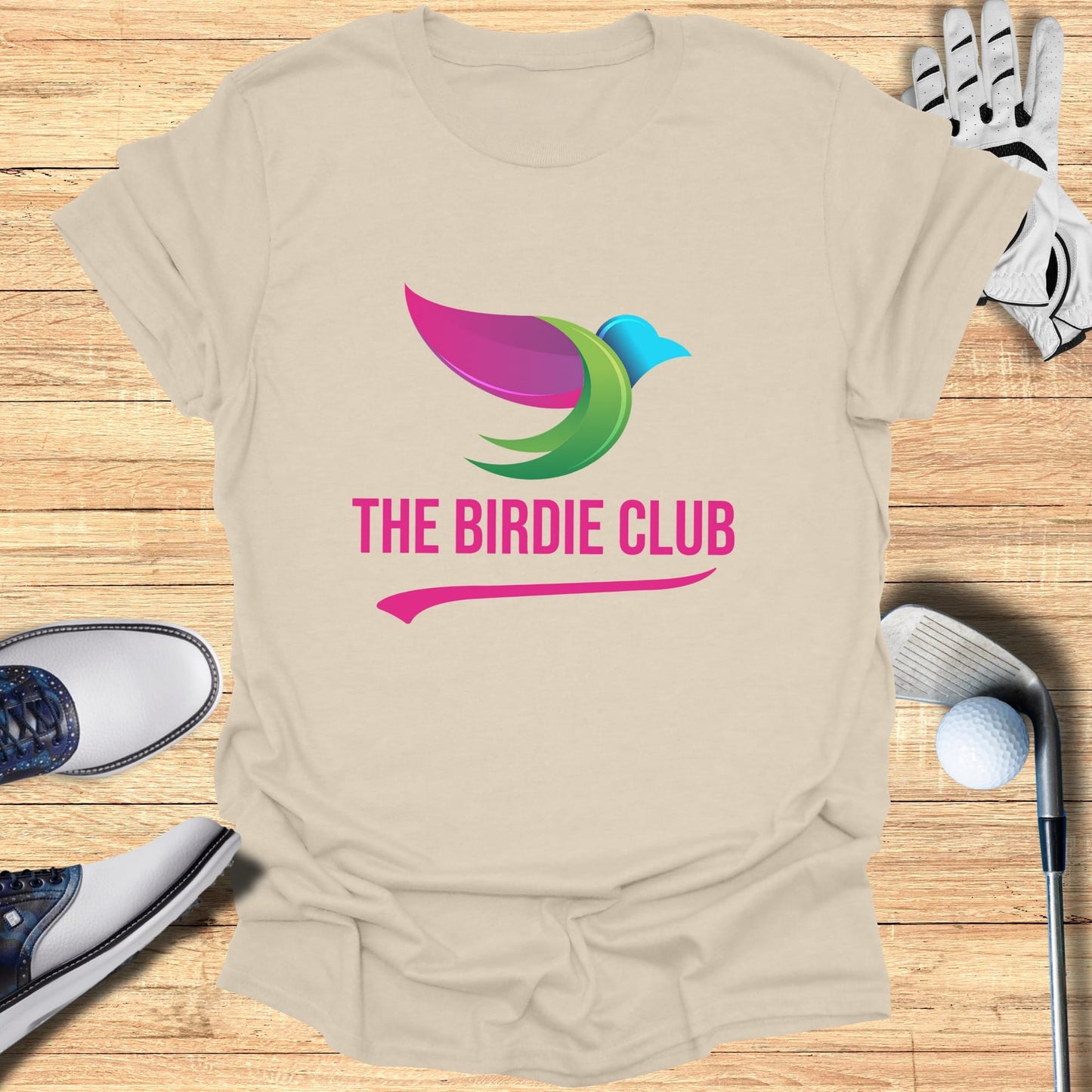 The Birdie Club logo on a beige t-shirt with golf accessories.