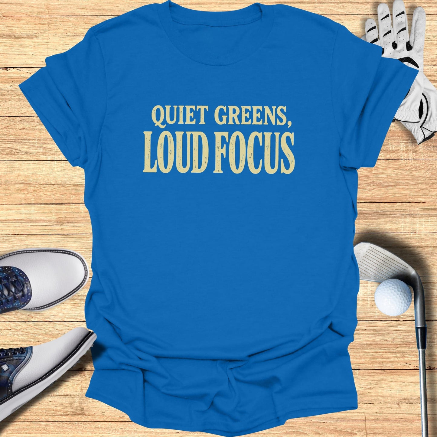Blue t-shirt with "QUIET GREENS, LOUD FOCUS" text, next to golf accessories.